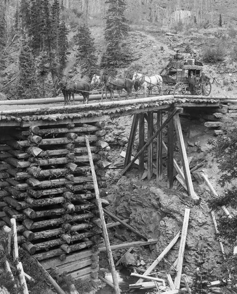 A stagecoach pulled by six horses crosses a rustic wooden bridge over a rocky ravine. The background features trees and a rugged landscape, suggesting a historical or pioneer setting.