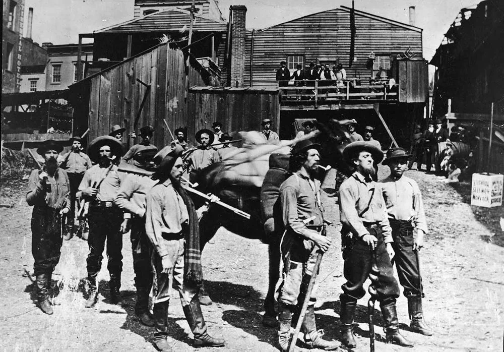 A group of men, dressed in late 19th to early 20th-century attire, stand outdoors in a dirt area. Some hold rifles. A wooden building is in the background, with more people visible on a balcony. The scene suggests a historical or military context.