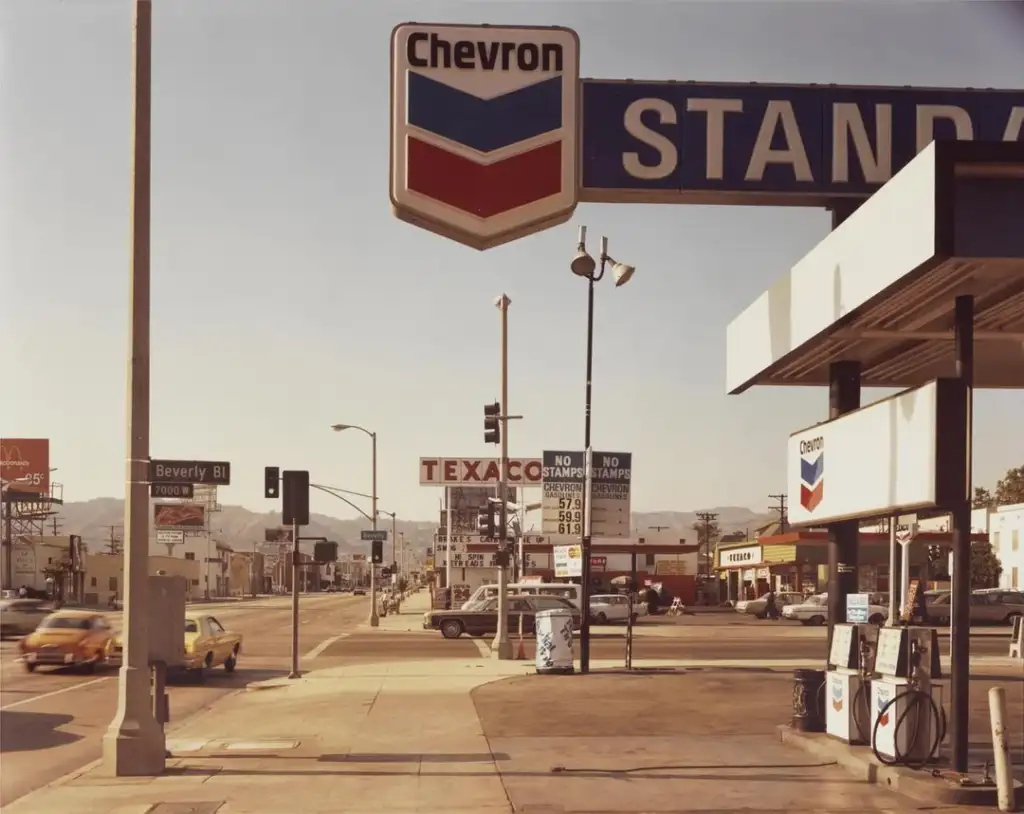 Vintage street scene featuring a Chevron gas station with a large sign and fuel pumps. The background shows a Texaco station, a road with traffic, and a McDonald's sign. The setting is urban, with hills visible in the distance under a clear sky.