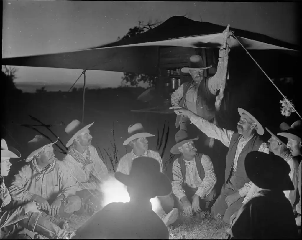 A group of cowboys sit around a campfire under a tarp at dusk, wearing hats and Western attire. One cowboy stands, holding a rope and pointing into the distance, while the others listen attentively. Shadows and the glow of the fire create a rustic ambiance.