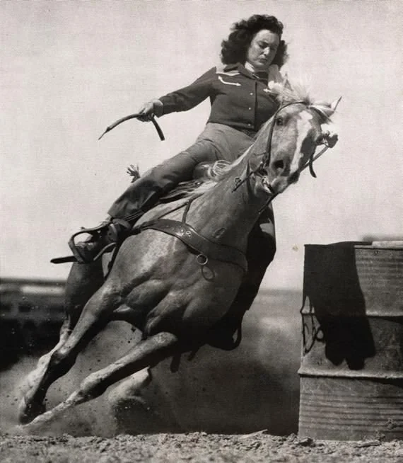 A woman in Western attire is riding a horse at high speed, rounding a barrel during a rodeo event. The horse is in mid-stride, illustrating the dynamic movement of barrel racing. The image is black and white, conveying a vintage feel.