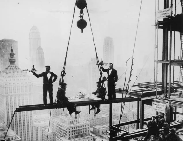 Workers having lunch on a steel beam high above New York City, with skyscrapers visible. Two waiters in tuxedos serve meals on trays. A table is set with dishes, creating a surreal, elegant dining scene in a construction setting.