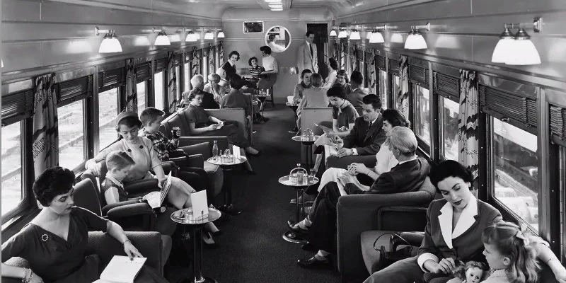 Black and white photo of a vintage train dining car. Men, women, and children are seated on upholstered chairs along both sides, reading or conversing. A waitress, visible near the center, serves a table. Large windows line the walls.