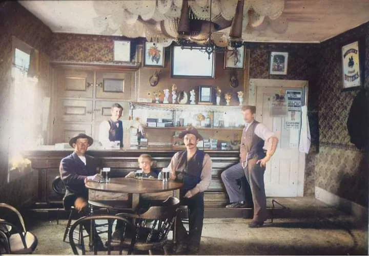 A vintage photo of five men in an old-fashioned bar. Three men sit at a round table with drinks, while two men stand behind them. The room features ornate wallpaper, a chandelier, and various items behind the bar counter.