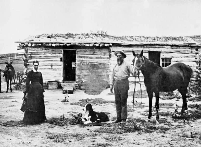 A black and white photo of a woman and a man standing in front of a log cabin. The man holds a horse, and there is a dog lying on the ground between them. Another horse is tied up in the background near the cabin.