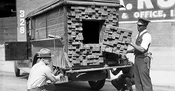 Two men inspect a truck with a hidden compartment filled with stacks of bricks. One man holds a brick panel. A third man crouches beneath the truck. The scene appears to be a historical investigation or inspection.