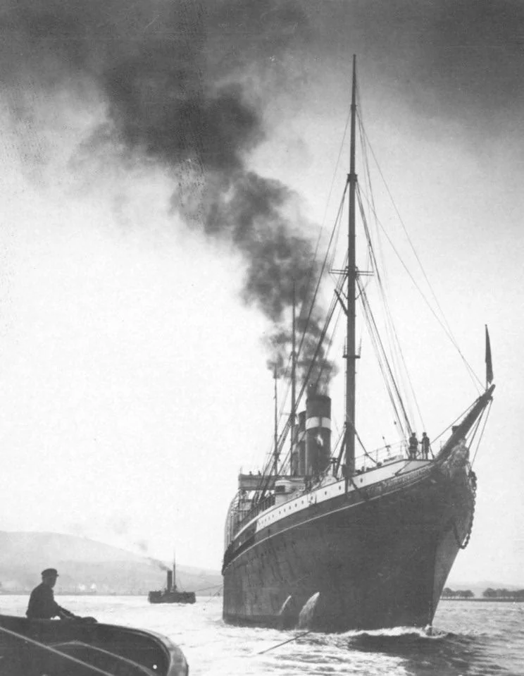 A large steamship emitting dark smoke sails through calm waters, viewed from a low angle. A small boat with a person sitting inside is in the foreground. Another distant vessel is seen against a backdrop of hills and a hazy sky.