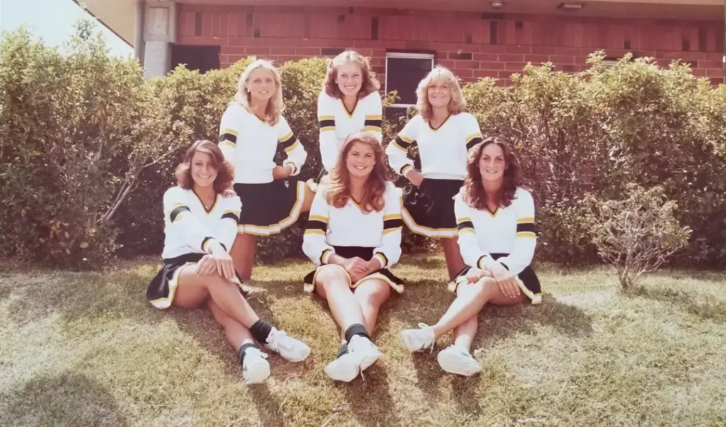 Six women in cheerleader uniforms sit and kneel on grass, posing for a group photo. They wear matching outfits with white tops, black skirts, and yellow accents. Bushes and a brick building are in the background.