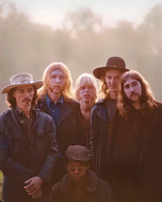 Six people are outdoors, dressed in 1970s-style clothing. Four men with long hair, some wearing hats, stand in the back. A woman with blond hair is in the middle, and a man with glasses and a flat cap sits in the front.