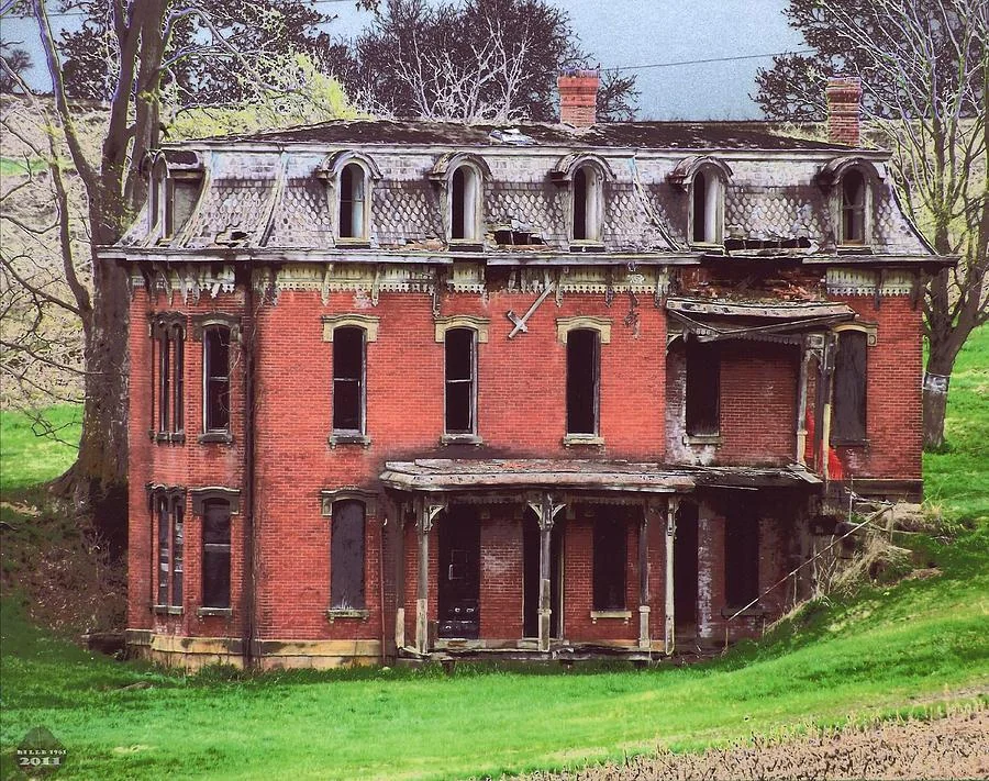 An abandoned, weathered red brick house with a partially collapsed roof and broken windows. The structure has ornate architectural details, including decorative trim and two chimneys. It's surrounded by grassy land and trees in the background.