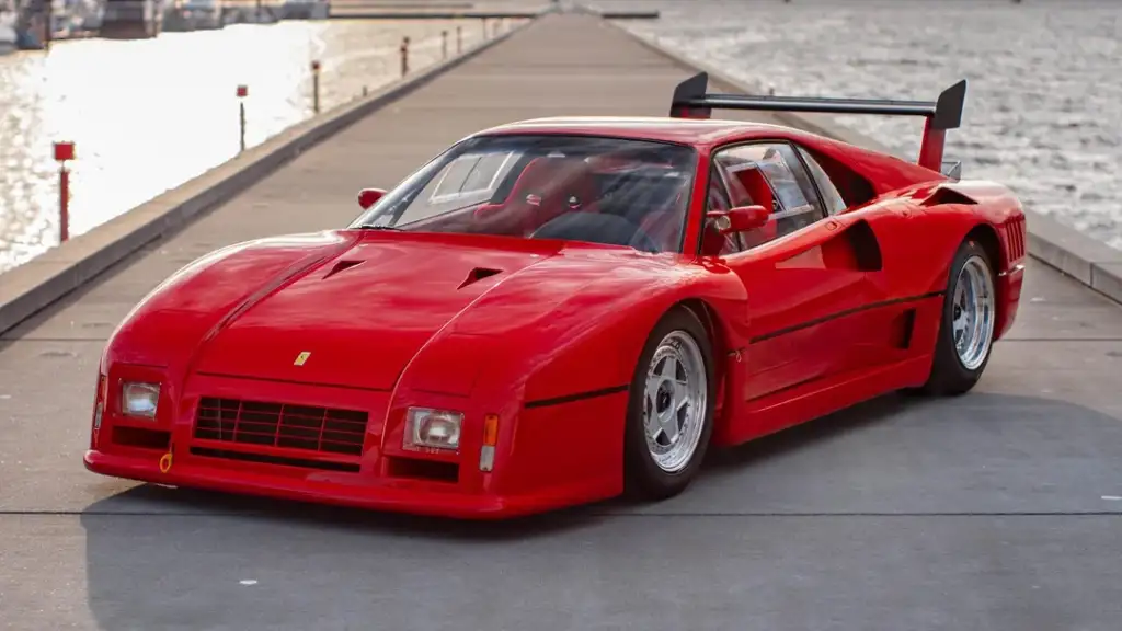 A sleek, red sports car with a rear spoiler is parked on a pier, with water on both sides. The hood has vents, and the car's design features sharp angles and a low profile. The sun is setting, casting a warm glow on the scene.