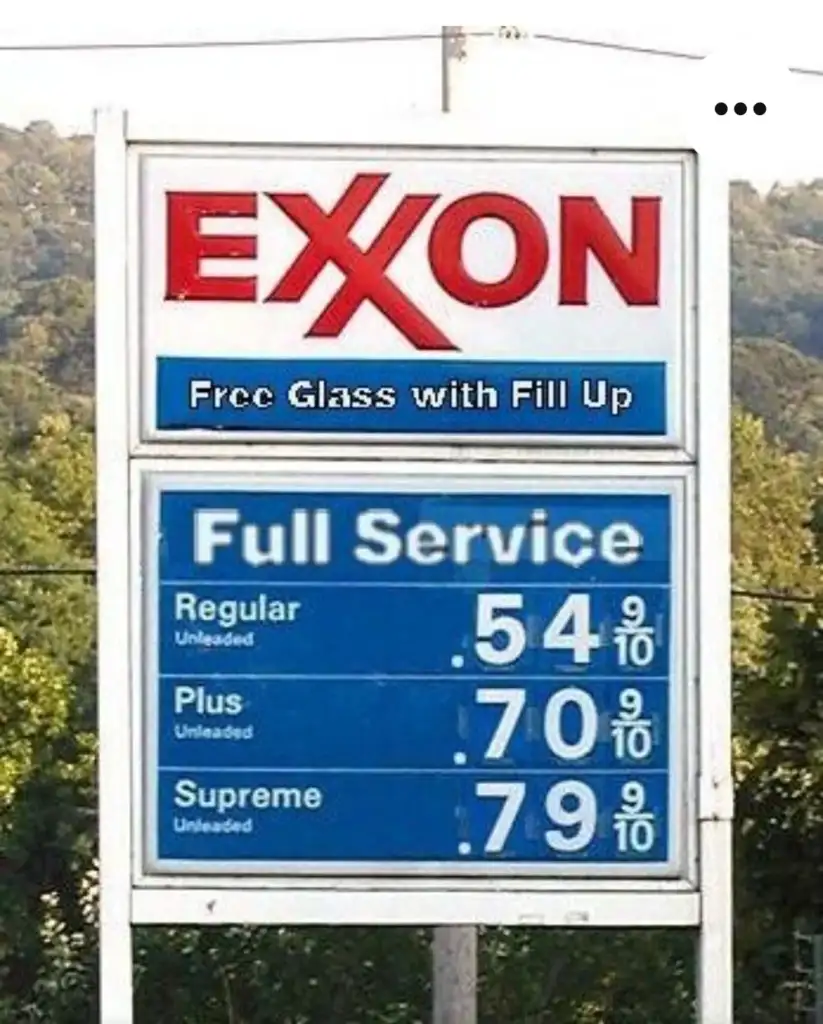 A vintage Exxon gas station sign displays prices: Regular Unleaded at 54.9 cents, Plus Unleaded at 70.9 cents, and Supreme Unleaded at 79.9 cents. The sign offers "Free Glass with Fill Up." Trees are visible in the background.