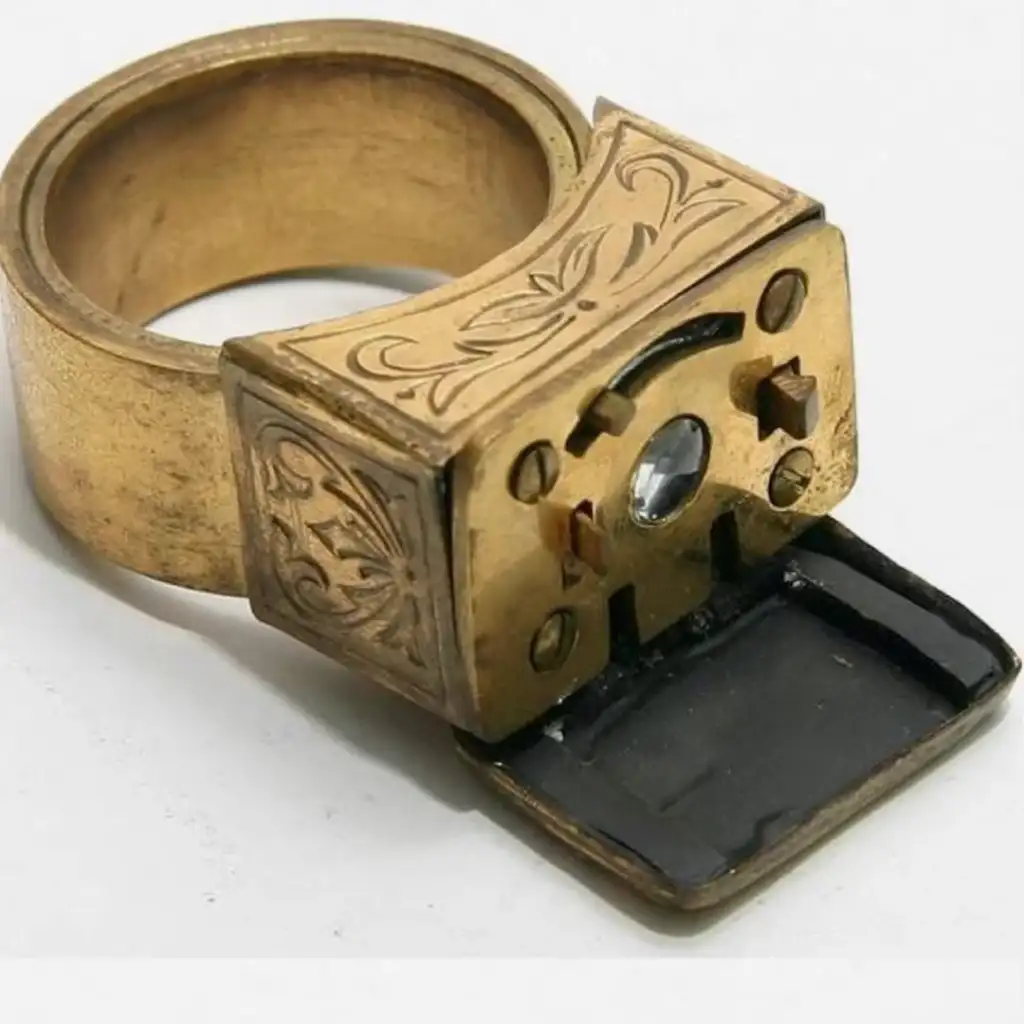 A vintage gold ring with an intricate engraved design features a small hidden compartment. The compartment is open, revealing an inner mechanism. The ring has an antique, ornate appearance.