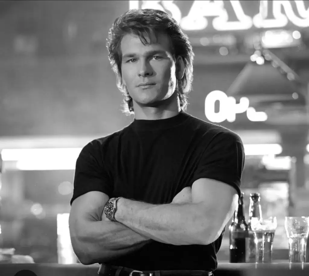 A person with wavy hair stands confidently with arms crossed in a bar setting. They wear a dark t-shirt and watch. Several drink bottles and glasses are on the table beside them. Bright lighting and neon signs are in the background. The image is in black and white.