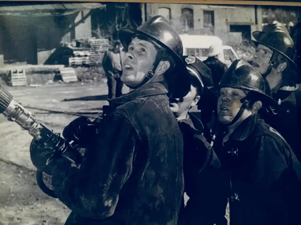 A group of firefighters in helmets and uniforms are looking upwards with focused expressions. One firefighter is holding a hose nozzle, ready for action. The background shows a partially visible building and some equipment. The image is in black and white.