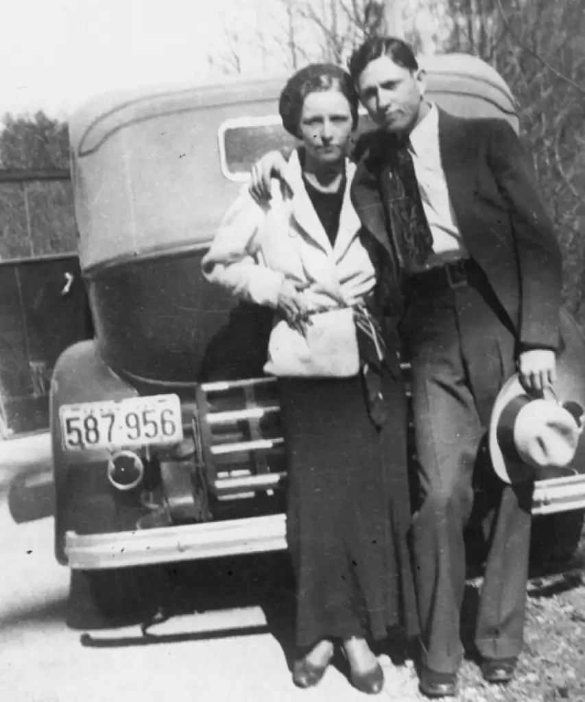 A black and white photo of a woman and a man leaning against an old-fashioned car. The woman is wearing a long skirt and a coat, while the man is in a suit, holding a hat. The car's license plate reads "587-956." They are outside in daylight.