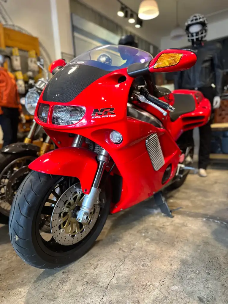 A bright red motorcycle with the letters "NR" on its side is displayed indoors. It has a sleek design, black seat, and shiny disc brakes, and stands on a concrete floor with apparel and accessories in the background.