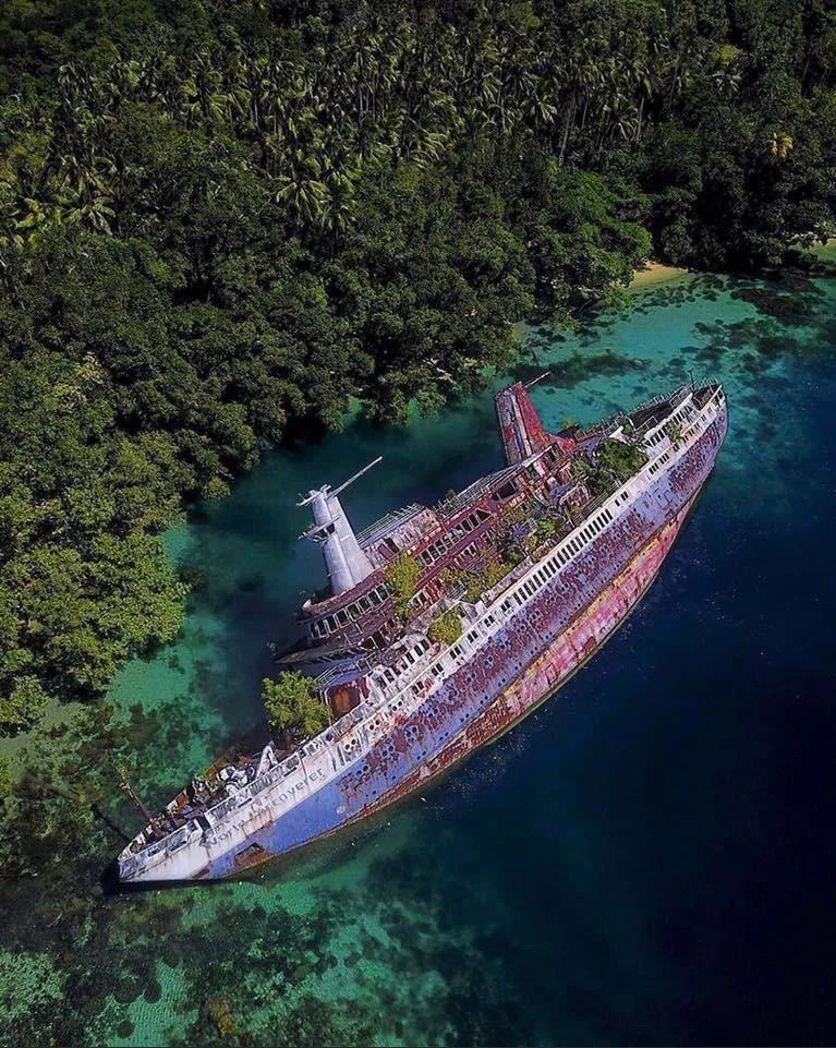 Aerial view of a large, rusted shipwreck partially submerged in clear turquoise waters. The vessel is surrounded by lush green forest, and the deck is overgrown with vegetation, indicating it has been abandoned for some time.