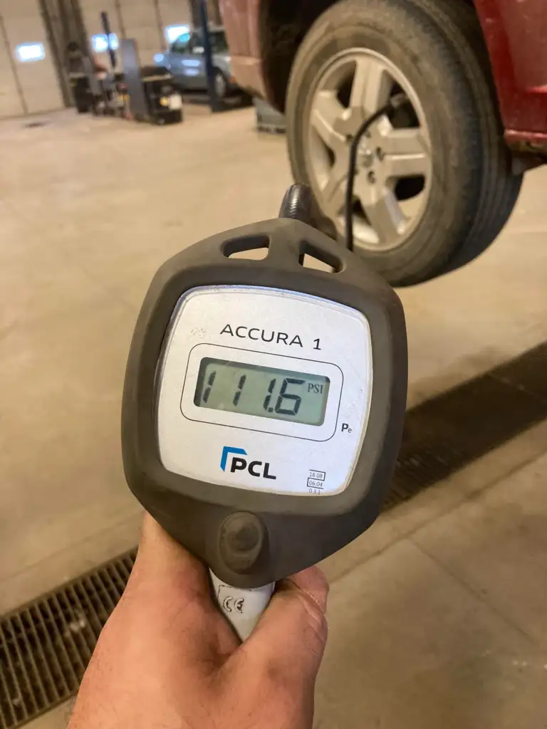 Close-up of a hand holding a tire pressure gauge reading 11.6 PSI. In the background, a red car is lifted with a visible wheel. The gauge is labeled "Accura 1" by PCL. The setting appears to be a garage or repair shop.