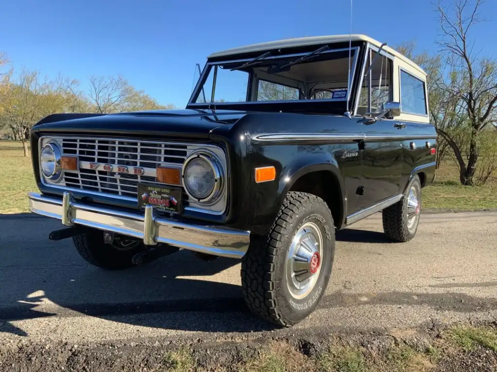 A classic black Ford Bronco with a white roof parked on a road. It features distinctive round headlights and a chrome grille. Trees and grass are visible in the background on a sunny day.