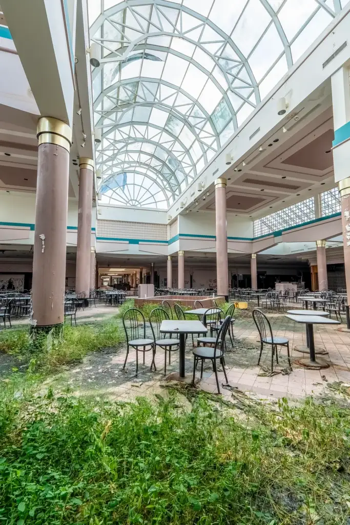 Deserted mall interior with overgrown plants covering the floor. Empty metal tables and chairs are scattered around. The ceiling features a large glass dome, allowing natural light in, highlighting the decay and abandonment.