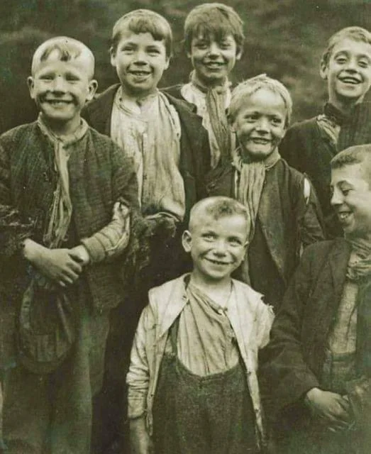 A vintage photo of seven young boys smiling and wearing worn, casual clothing. They stand closely together, some with hands in pockets. The background is blurred, focusing attention on their joyful expressions.