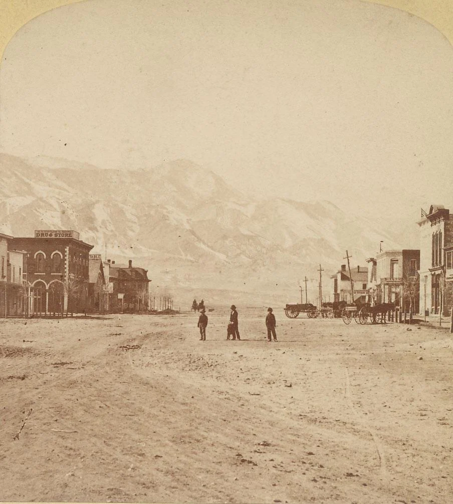 A sepia-toned photograph of a dusty street in a small 19th-century town. Three people stand in the center with mountains in the background. On either side, wooden buildings line the street, and a horse-drawn wagon is visible on the right.