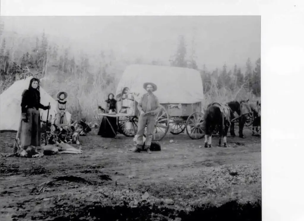 A vintage photo shows a man with a hat standing in front of a covered wagon. Two women and a child are on the left, with a campfire nearby. Two horses are hitched to the wagon, surrounded by trees and a barren landscape.