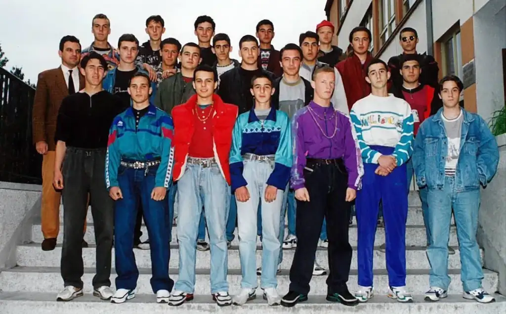 A group of young men and a teacher stand on stairs for a group photo. They are wearing casual 90s attire, including colorful jackets, jeans, and sneakers. The setting looks like a school or campus with buildings in the background.