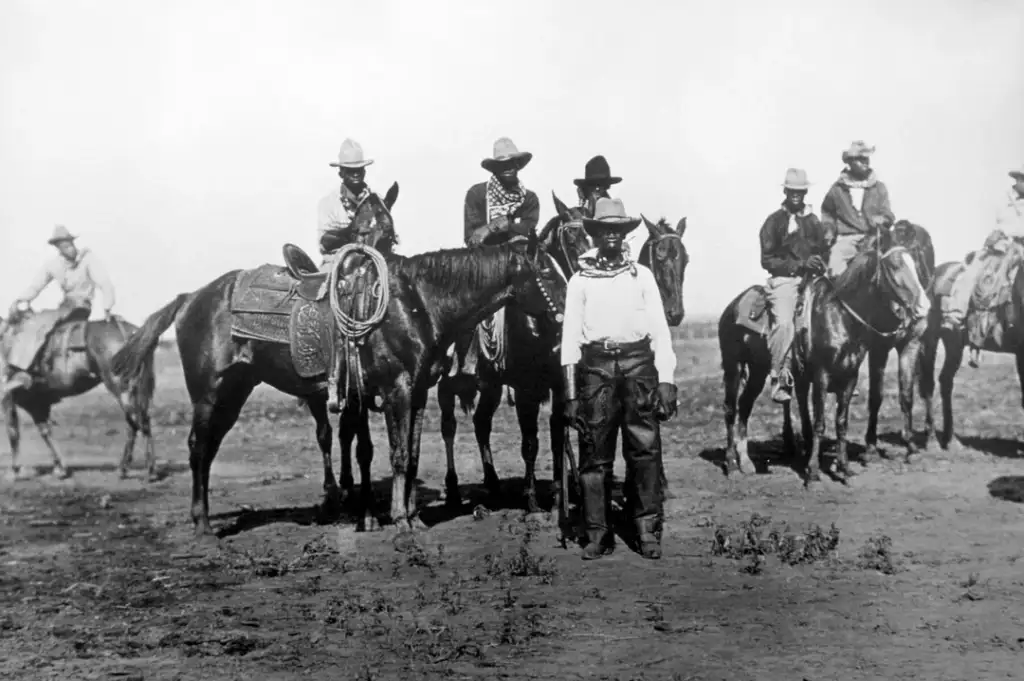 A historical black and white photo of seven cowboys, six on horseback and one standing beside a horse, in an open landscape. They wear wide-brimmed hats and bandanas, showcasing traditional Western attire.