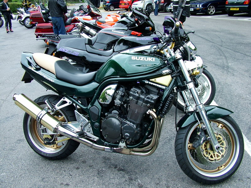 A green Suzuki motorcycle with exposed engine parts, parked among other bikes in a lot. The bike features gold accents on the rims and a metallic exhaust pipe. Other motorcycles and a car are visible in the background.