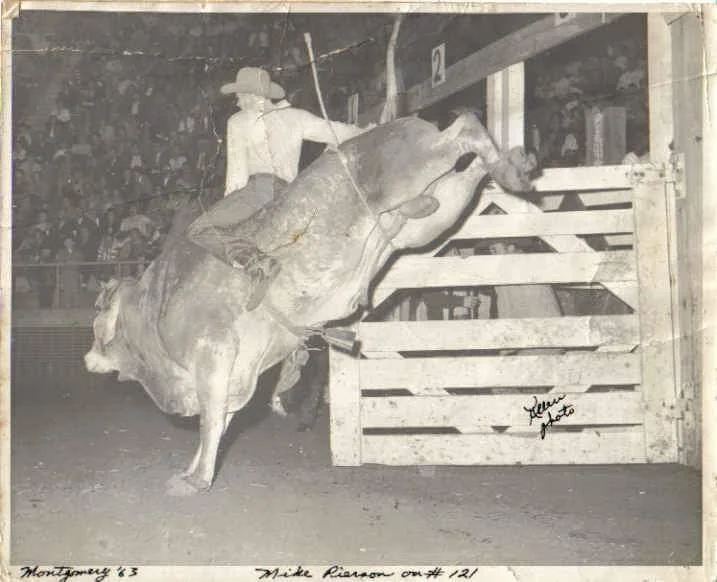 A rodeo cowboy rides a bucking bull, holding onto a rope with one hand. The cowboy wears a wide-brimmed hat and boots. The scene takes place in an arena with a visible audience in the background. The image is in black and white.