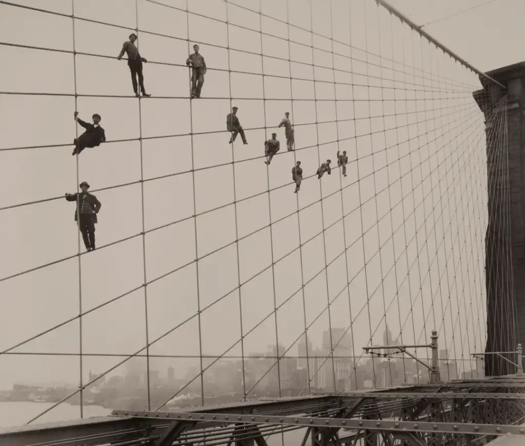 Workers climb and balance on the suspension cables of a large bridge under construction. The skyline is visible in the background, showing a distant city view.