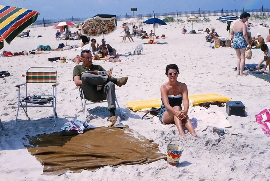 A man sits on a striped chair reading a newspaper, while a woman in sunglasses sits on a yellow floating mat on a sandy beach. They are surrounded by beachgoers, umbrellas, and towels under a clear blue sky.