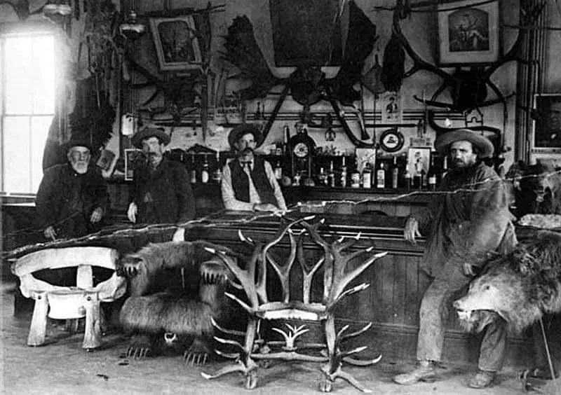 Black and white photo of four men in a bar decorated with hunting trophies. The men stand behind the bar, which features bear rugs and antler chairs. Various bottles and items are on the bar, and taxidermy mounts adorn the walls.