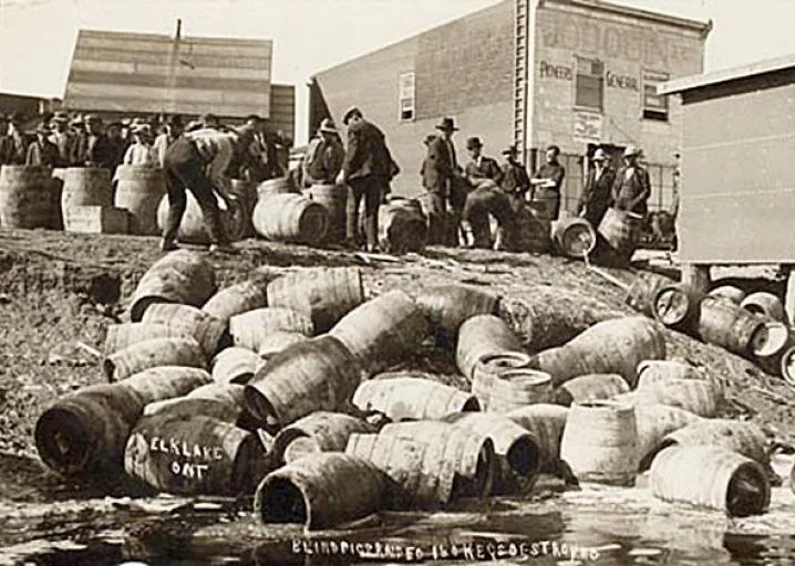 A group of people stands on a dirt slope surrounded by numerous barrels. Some individuals are interacting with the barrels, which are scattered around the area. Buildings in the background have visible signage. The scene appears historical.