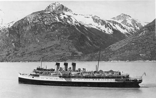 A black and white photo of a large ocean liner with multiple smokestacks sailing through calm waters. Snow-capped mountains rise in the background under a clear sky.