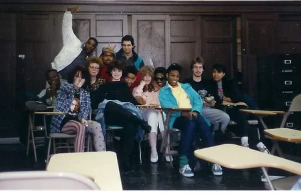 A group of people sitting and standing around desks in a classroom. They are casually dressed, and some are smiling, while one person in the back raises an arm. The room has wooden paneling and filing cabinets in the background.