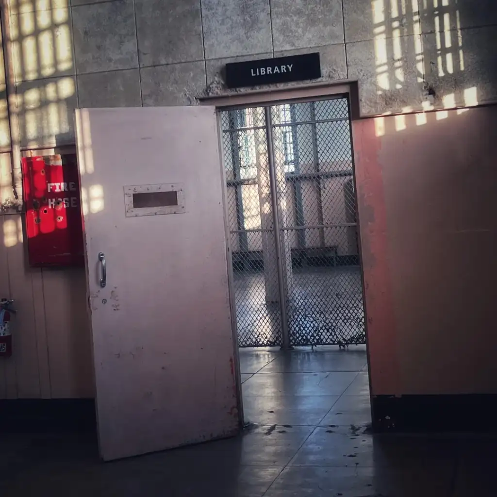 A dimly lit hallway with a sign above a doorframe labeled "Library." The metal door is ajar, leading to a barred gate. Light filters through high windows, casting shadows on the worn walls. A red fire hose cabinet is mounted on the left wall.