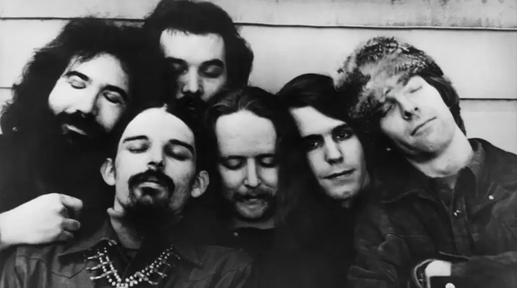 A black and white photo of six men standing closely together in front of a wooden wall. They have long hair and varied facial hair styles. The man on the right is wearing a furry hat with his eyes closed, while the others are looking forward or down.