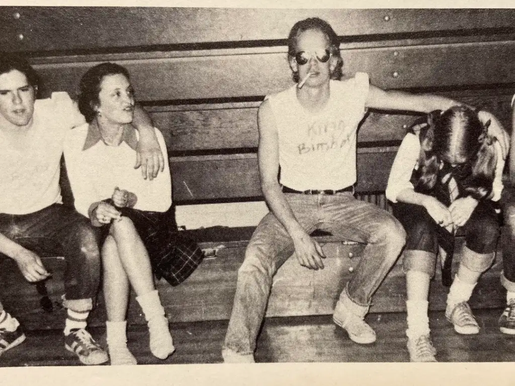 A group of four people sitting on wooden bleachers. One person is wearing sunglasses and a sleeveless shirt, smoking a cigarette. Another person leans forward with their head down. The others are casually seated, engaging in conversation.