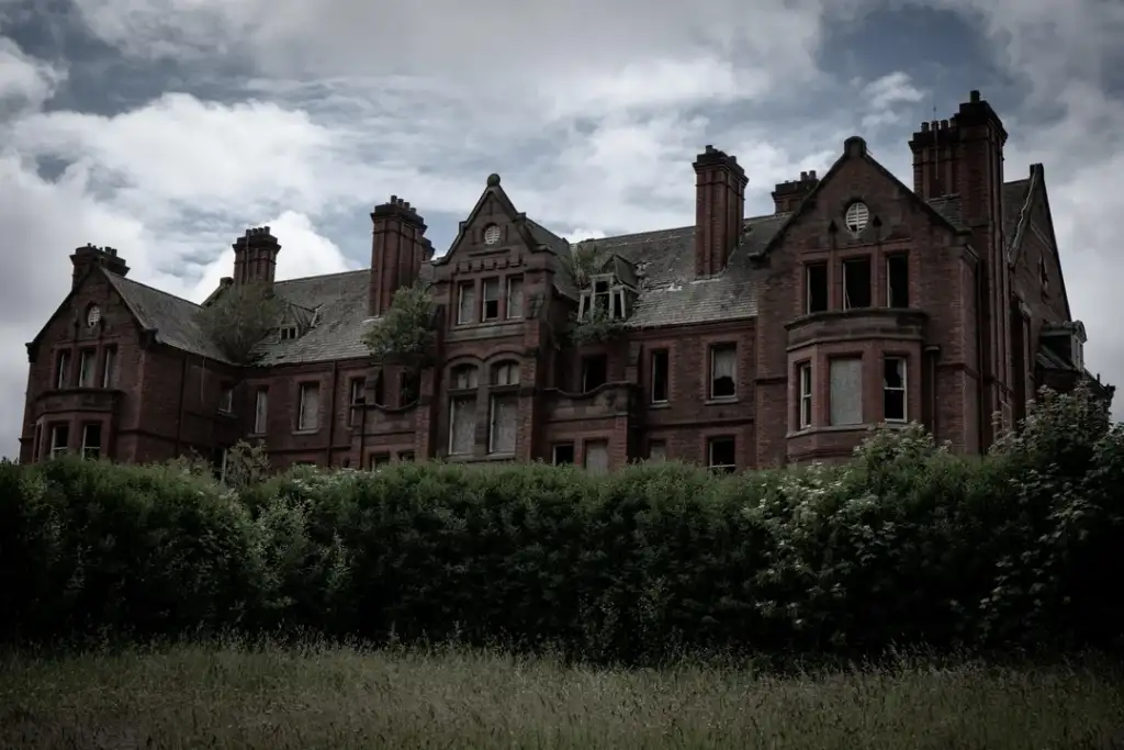 A large, abandoned brick mansion stands behind overgrown bushes under a cloudy sky. The building is weathered, with visible broken windows and vegetation growing on the roof, creating an eerie and desolate atmosphere.