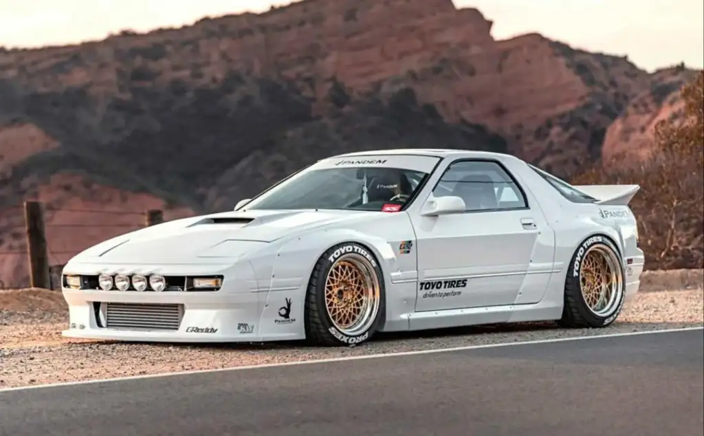 A modified white sports car is parked on a road with scenic rocky hills in the background. The car features a wide body kit, gold mesh wheels, and various decals. The setting is outdoors, during the day.