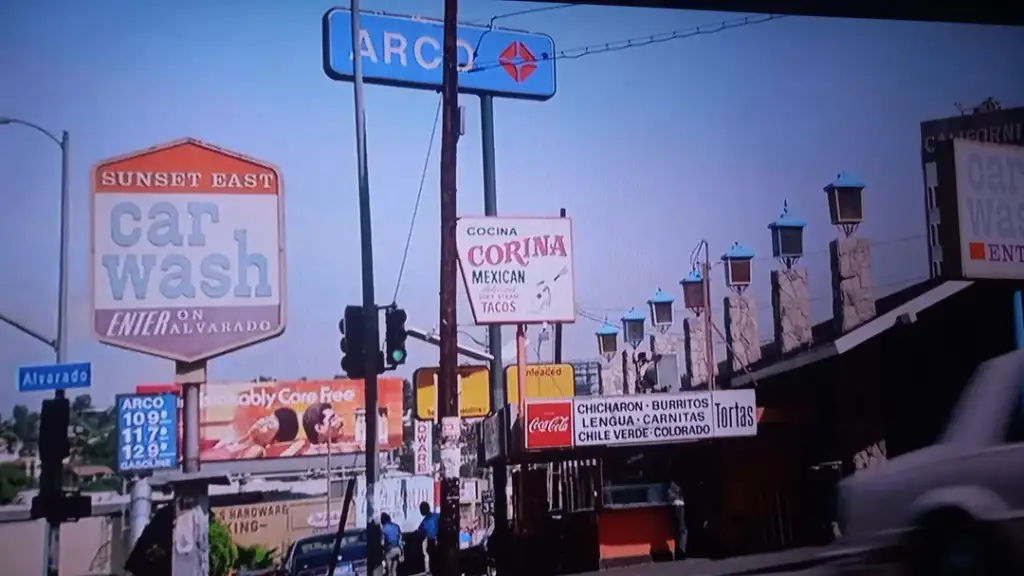 Street scene featuring several signs, including ARCO gas prices and Sunset East Car Wash. The image also shows signs for Cocina Corina Mexican Tacos and other eateries. The scene includes traffic lights and a partially visible car.