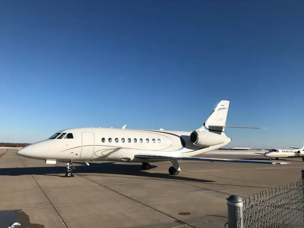 A sleek private jet with white livery is parked on a sunlit tarmac under a clear blue sky. In the background, another smaller aircraft can be seen. A chain-link fence runs along the right side of the image.