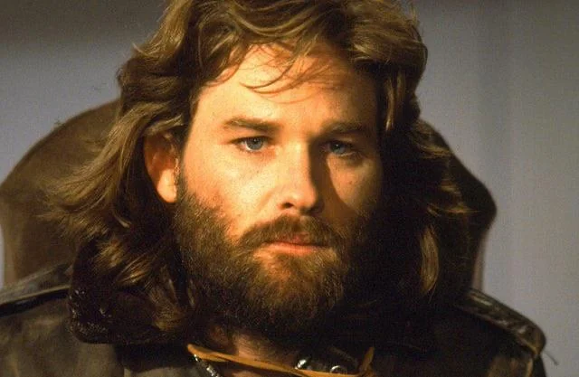 A person with long, wavy hair and a full beard is looking intently forward. They are wearing a dark leather jacket and are slightly illuminated, creating a focused and thoughtful expression.