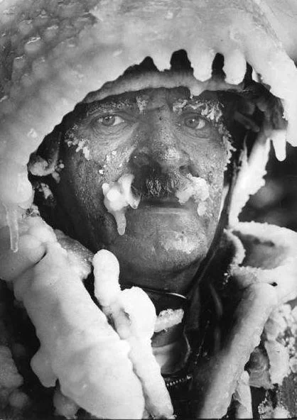 A person wearing a heavily iced coat and hood, with their face partially covered in ice. Their expression is serious, and the icy buildup gives a dramatic depiction of extreme cold weather conditions.