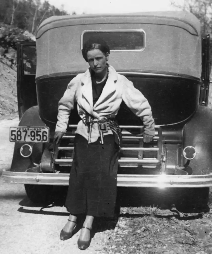 A black and white photo shows a woman leaning against the front of a vintage car. She is wearing a light-colored jacket and dark skirt, with a belt around her waist. The car's license plate reads "587-956." The scene appears to be outdoors.