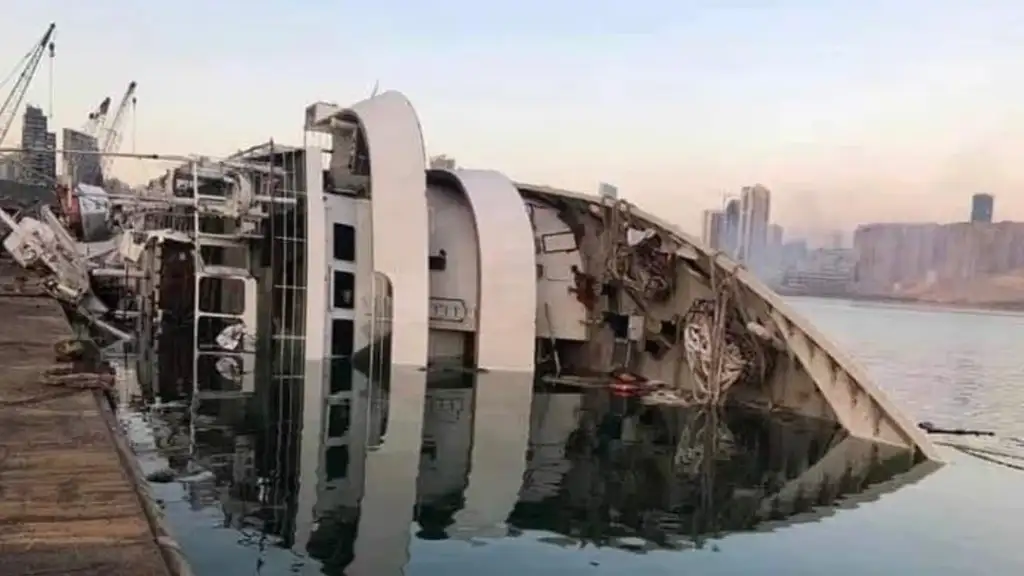 A large ship is partially submerged and tilted on its side near a dock. The city skyline and calm water reflect light in the background. The ship shows visible damage and debris is scattered around the site.