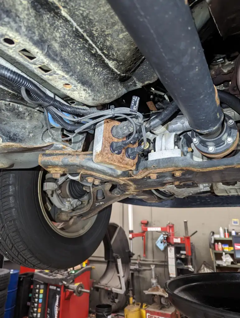 View of a car's undercarriage showing a suspension component with visible rust. A large black pipe runs across the image. A tire is on the left and various tools and equipment are in the background, suggesting a workshop setting.